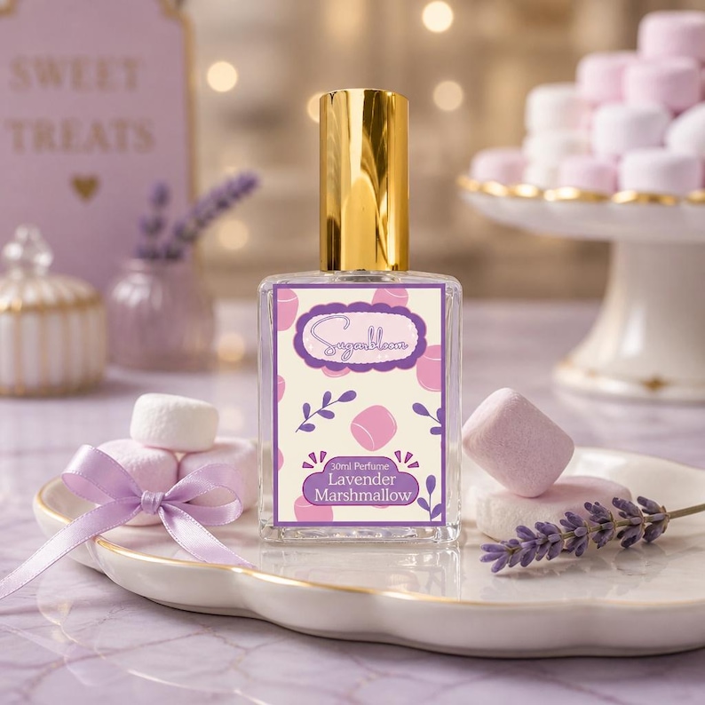 May include: A perfume bottle with a gold cap and a label that reads "Lavender Marshmallow." The label has a light purple background with pink marshmallow illustrations. The bottle sits on a white plate with marshmallows and a lavender sprig.