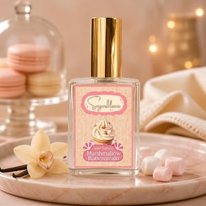 Puede incluir: Un frasco de perfume transparente con tapa dorada, etiquetado "Sugarbloom" y "Marshmallow Buttercream". La etiqueta presenta una ilustración de cupcake. La imagen incluye macarons, malvaviscos y una flor de vainilla, lo que sugiere un tema de fragancia dulce.