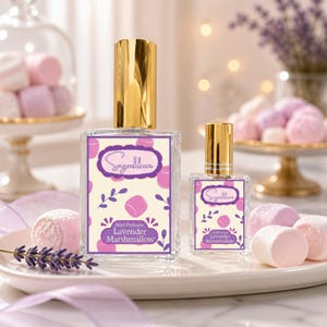 May include: Two perfume bottles with gold caps and labels that read "Sugarbloom Lavender Marshmallow." The labels feature a purple design with marshmallow and leaf illustrations. The bottles are on a white plate with marshmallows and lavender sprigs.