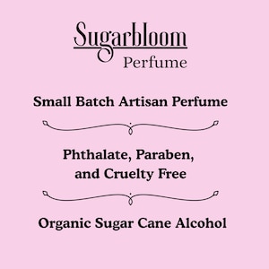 May include: A pink background with the text "Sugarbloom Perfume" in a decorative font. Below, it reads "Small Batch Artisan Perfume," followed by "Phthalate, Paraben, and Cruelty Free," and "Organic Sugar Cane Alcohol."