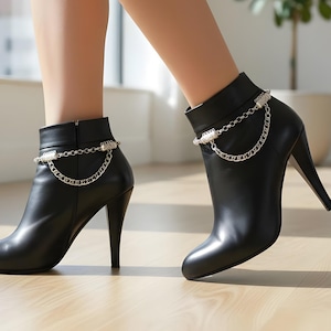 May include: Black ankle boots with stiletto heels and a silver chain detail. The boots have a smooth, polished finish and a zip closure. The heels are high, and the boots are suitable for formal or casual wear. The boots are made of a smooth material.