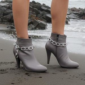 May include: Gray ankle boots with high heels and silver chain detailing. The boots have a zipper closure and are worn with denim shorts. The image is taken on a beach with a rocky shoreline and a misty background.
