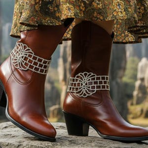 May include: Brown leather cowboy boots with decorative rhinestone ankle cuffs. The boots have a pointed toe, a block heel, and a mid-calf height. The boots are worn with a floral print dress.
