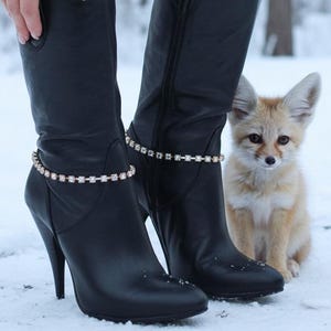 May include: Black leather high-heeled boots with a decorative rhinestone ankle bracelet. The boots have a pointed toe and a high heel. A small fox with large ears sits in the snow.