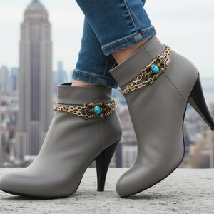 Peut inclure: Bottines grises à talons noirs, ornées de chaînes dorées avec des détails turquoise. Ces bottines au design moderne sont portées avec un jean bleu, sur fond de paysage urbain.