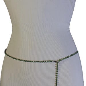 K&ouml;nnte beinhalten: Eine zarte gr&uuml;ne Strass-Taillenkette. Die Kette wird auf einer wei&szlig;en Schaufensterpuppe pr&auml;sentiert, um ihr Design zu zeigen. Sie besteht aus kleinen, runden gr&uuml;nen Strasssteinen, die durch d&uuml;nnes, silberfarbenes Metall verbunden sind.