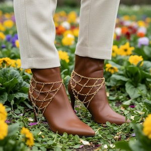 May include: Brown leather ankle boots with pointed toes and high heels. The boots are adorned with gold chain anklets. The person is wearing beige pants. The background is a flower bed with yellow and purple flowers.