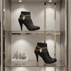 Könnte beinhalten: Schwarze Stiefeletten mit goldenen Ketten und Kristallakzenten. Die Stiefel haben eine spitze Zehenpartie und einen hohen, geschwungenen Absatz. Die Stiefel werden auf einem Glasregal in einem gut beleuchteten Schrank ausgestellt.