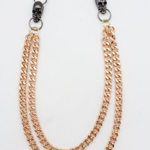 May include: A gold-colored chain with two parallel strands, featuring black skull accents and a gold-tone clasp and ring. This accessory is designed to be attached to a belt loop.