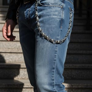 Può includere: Una catena color argento con maglie a forma di teschio pende dal passante dei jeans blu. La catena è attaccata a una cintura di pelle nera. È visibile una giacca di pelle nera.