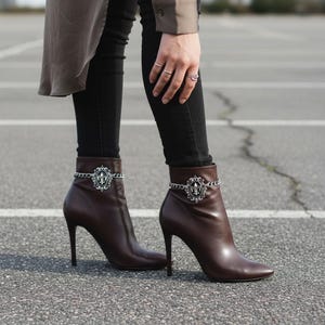 Könnte beinhalten: Braune Leder-Ankle Boots mit hohen Absätzen. Jeder Stiefel hat eine dekorative silberne Kette und ein Fleur-de-Lis-Emblem. Die Stiefel werden mit einer schwarzen Hose getragen.