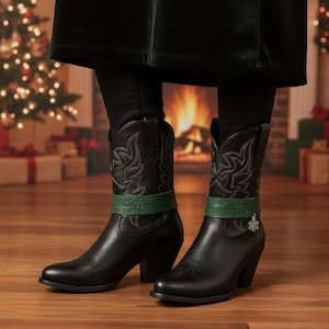 May include: Black leather cowboy boots with decorative stitching and a green band. The boots have a pointed toe and a stacked heel. A small charm hangs from the green band on one boot. The boots are on a wooden floor.