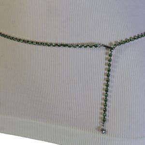 K&ouml;nnte beinhalten: Ein zarter, gr&uuml;ner Strass-Tailleng&uuml;rtel. Der G&uuml;rtel besteht aus kleinen, runden, gr&uuml;nen Strasssteinen, die zu einer Kette aufgereiht sind. Ein vertikaler Strang aus Strasssteinen h&auml;ngt von der Mitte herab und endet in einem kleinen, silberfarbenen Anh&auml;nger.