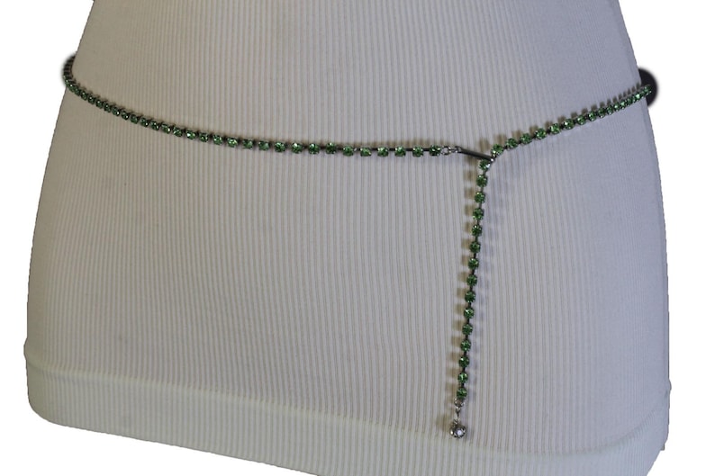K&ouml;nnte beinhalten: Ein zarter, gr&uuml;ner Strass-Tailleng&uuml;rtel. Der G&uuml;rtel besteht aus kleinen, runden, gr&uuml;nen Strasssteinen, die zu einer Kette aufgereiht sind. Ein vertikaler Strang aus Strasssteinen h&auml;ngt von der Mitte herab und endet in einem kleinen, silberfarbenen Anh&auml;nger.