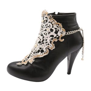 Puede incluir: Bota negra de tacón con una superposición de encaje blanco y una cadena plateada con un dije de perla.