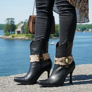 May include: Black leather heeled boots with pointed toes and decorative lace and gold accents. The boots have a side zipper and are worn with black jeans. A small brown purse hangs in the background.