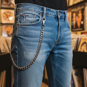 Puede incluir: Vaqueros de mezclilla azul claro con un accesorio de cadena plateada para la cartera. Los vaqueros tienen un diseño clásico de cinco bolsillos y un lavado medio. La cadena plateada está sujeta al trabilla y al bolsillo, añadiendo un detalle elegante. El fondo muestra estanterías de discos de vinilo.