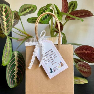 May include: A brown paper gift bag with a white lace ribbon and a thank you tag. The tag has a pink and white checkered background with a cartoon illustration of dogs and the text "Thank you for coming to my birthday party! I'm grateful to have a friend in you! - Micah".