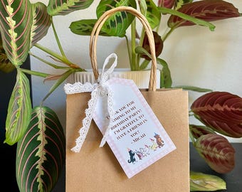 May include: A brown paper gift bag with a white lace ribbon and a thank you tag. The tag has a pink and white checkered background with a cartoon illustration of dogs and the text "Thank you for coming to my birthday party! I'm grateful to have a friend in you! - Micah".