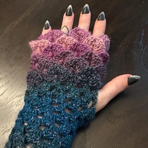 May include: A crocheted fingerless glove in shades of purple, blue, and teal, with a shimmering thread woven throughout. The glove features a scalloped design on the hand and a lacy pattern on the arm cuff. The cuff is approximately 6 inches long.