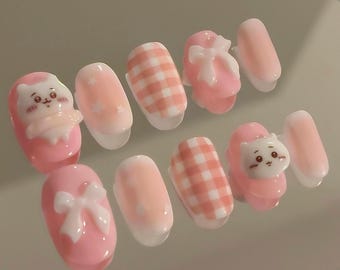 May include: A collection of ten artificial nails in a range of designs. The nails are primarily light pink with white details, such as bows, gingham patterns, and small cartoon cat and bear figures. The nails are oval-shaped.