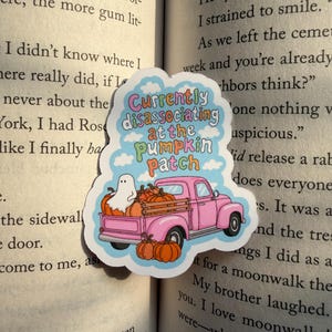 May include: A die-cut sticker featuring a pink vintage pickup truck loaded with orange pumpkins and a small white ghost. The text reads Currently disassociating at the pumpkin patch in colorful bubble letters against a light blue sky with white clouds.