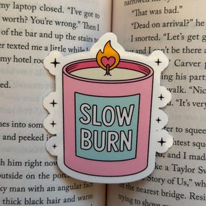 May include: A die-cut sticker featuring a pink candle in a jar with a light blue label that reads SLOW BURN in white capital letters. The candle has a flame with a small pink heart inside and is surrounded by small black four-pointed star icons.