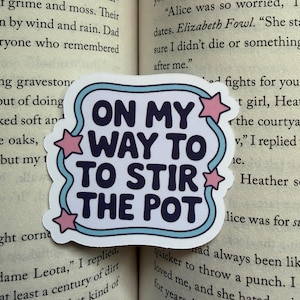 May include: A die-cut sticker featuring the dark blue text ON MY WAY TO STIR THE POT in a bold, rounded font. The text is enclosed by a light blue wavy border and decorated with four small pink stars positioned along the edges of the frame.