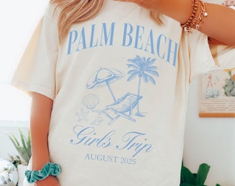 May include: A white t-shirt with a blue graphic design that says "Palm Beach Girls Trip August 2025". The graphic includes a palm tree, beach chair, and umbrella.