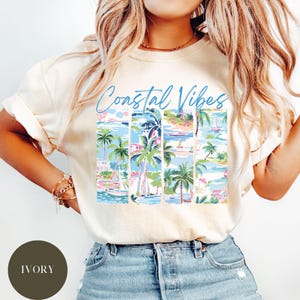 May include: A cream colored t-shirt with the text "Coastal Vibes" in blue script. The shirt features a graphic of palm trees, boats, and beach scenes in a vintage style.