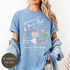 May include: Light blue t-shirt with the text "Savannah Bach Club EST 2026" and watercolor illustrations of a sailboat, building, and peaches. The text "Lauren's Bachelorette Savannah, GA" is also printed on the shirt. The shirt is made of washed denim.