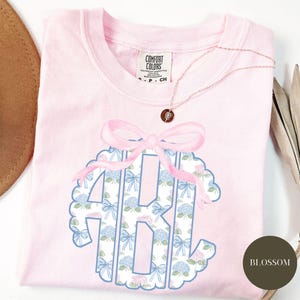 Op de afbeelding: Een lichtroze t-shirt met een groot monogramontwerp. Het monogram bevat de letters "ABI" in een bloemenpatroon met blauwe strikken en hortensia's. Bovenop het monogram zit een roze strik. Een gouden ketting is over het shirt gedrapeerd.