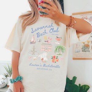 May include: Cream-colored t-shirt with a light blue graphic that reads "Savannah Bach Club" and "Lauren's Bachelorette, Savannah, GA." The graphic includes illustrations of a sailboat, cocktails, and a historic building.