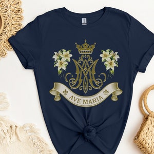 Puede incluir: Camiseta azul marino con un dise&ntilde;o dorado de Ave Mar&iacute;a que incluye una corona, lirios y un monograma.