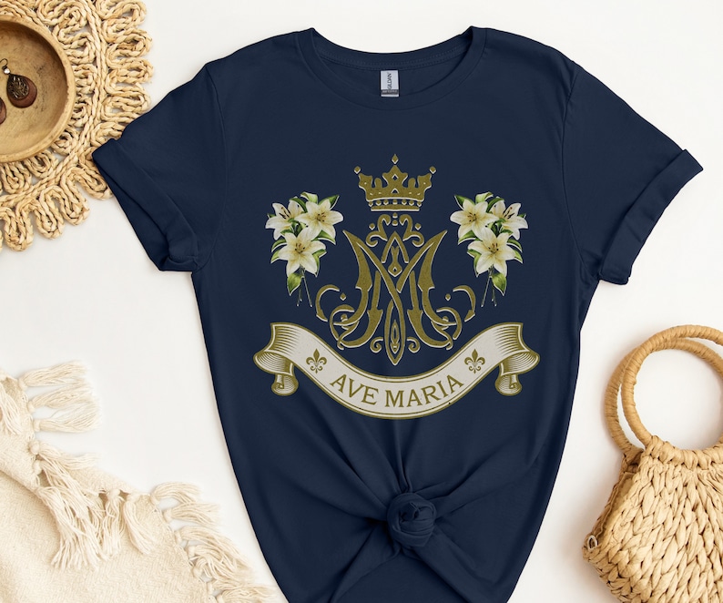 Puede incluir: Camiseta azul marino con un dise&ntilde;o dorado de Ave Mar&iacute;a que incluye una corona, lirios y un monograma.