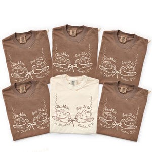 May include: A collection of six t-shirts, primarily in brown and one in cream, with a graphic design featuring cowboy hats and the text "Reckless in Texas, Aug 2026, Austin, TX." Each shirt has a small tag and a necklace.