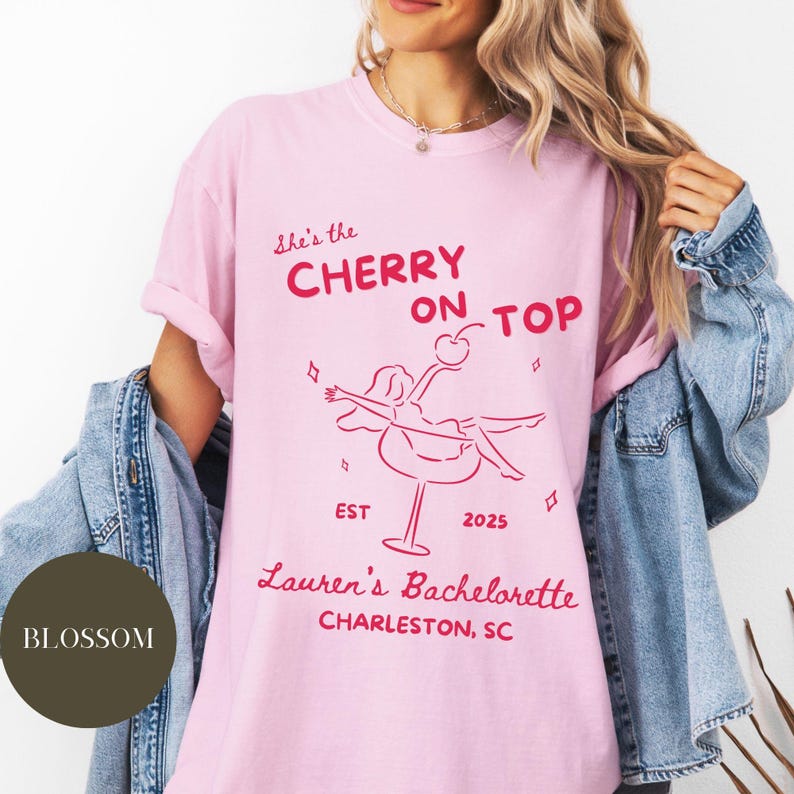 May include: Pink t-shirt with the text "She's the Cherry on Top" in red, along with a graphic of a woman in a cocktail glass. The text "Lauren's Bachelorette Charleston, SC" is also printed on the shirt.