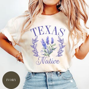 以下が含まれることがあります： 紫色の文字で「TEXAS Native」と書かれた白いTシャツ。緑の葉の輪が、紫色の野花の群れを囲んでいます。