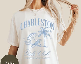 May include: Ivory-colored t-shirt with a light blue graphic that says "Charleston Girls Club" with a palm tree, beach chair, and cocktail design. The text "Lauren's Bachelorette" is also included. The shirt is a relaxed fit.