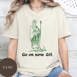 May include: A white t-shirt with a green illustration of a saint holding a staff. The text "Go on now. Git." is printed below the illustration.