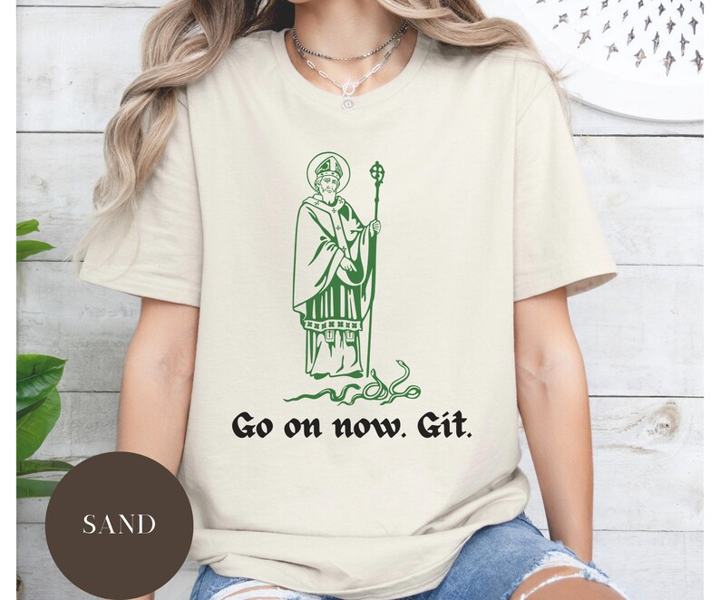 May include: A white t-shirt with a green illustration of a saint holding a staff. The text "Go on now. Git." is printed below the illustration.
