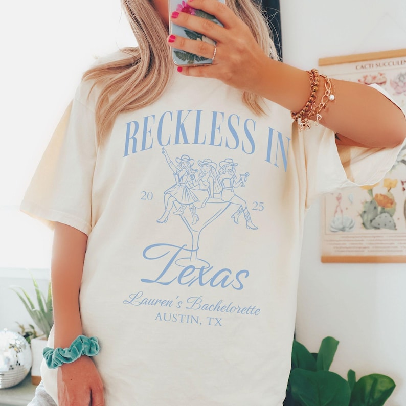 K&ouml;nnte beinhalten: Ein wei&szlig;es T-Shirt mit einem blauen Grafikdesign, auf dem "Reckless in Texas" steht, mit drei Frauen, die auf einem Martini-Glas sitzen. Die Grafik enth&auml;lt auch den Text "Lauren's Bachelorette Austin, TX" und die Zahlen "20" und "25".