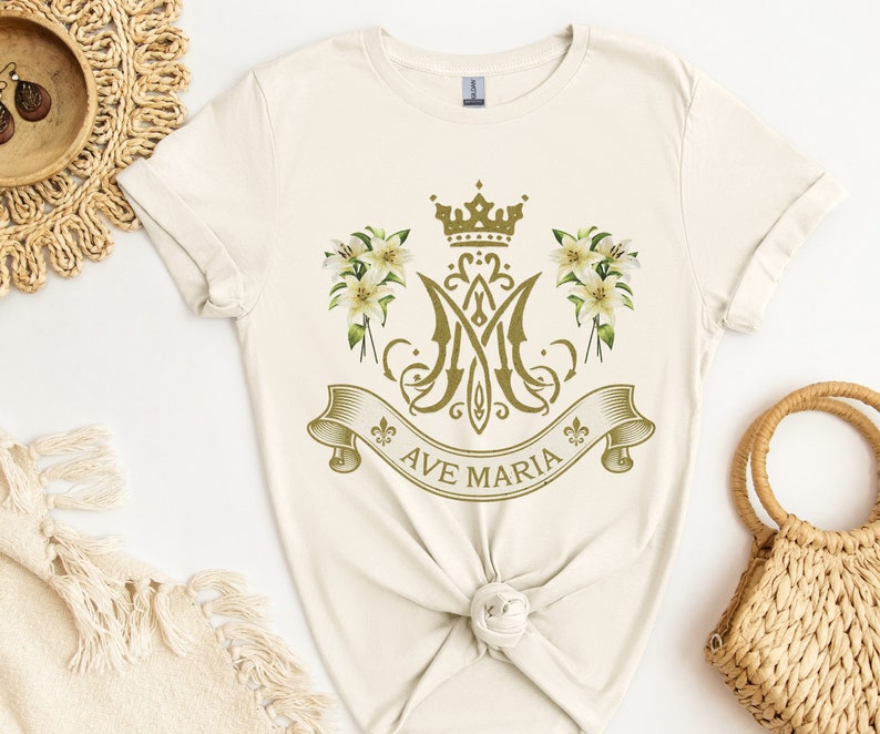 Puede incluir: Una camiseta de color crema con un gr&aacute;fico dorado de la Virgen Mar&iacute;a. El gr&aacute;fico presenta una corona, lirios y el texto "Ave Maria".