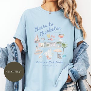 K&ouml;nnte beinhalten: Hellblaues T-Shirt mit einem grafischen Design einer Stadtlandschaft und dem Text "Cheers to Charleston est. 2023 Lauren's Bachelorette Charleston, SC".