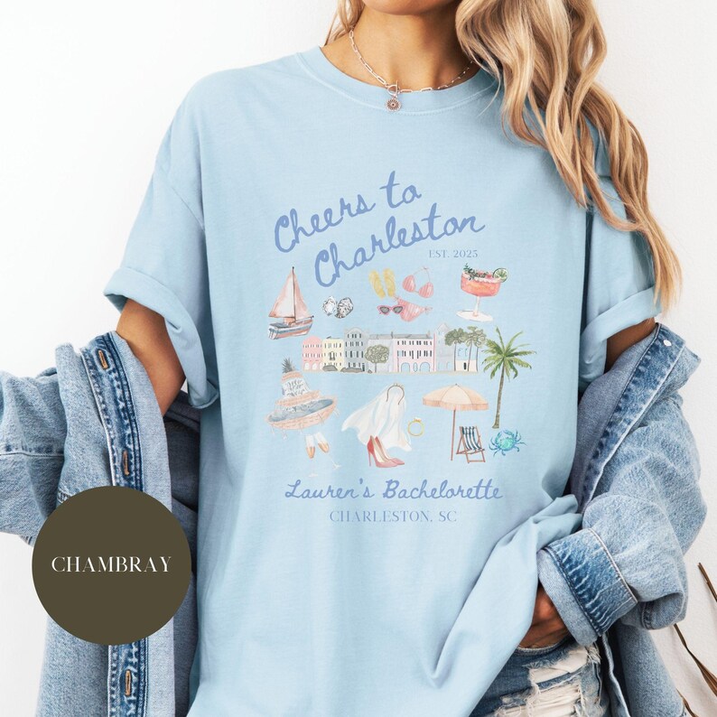 K&ouml;nnte beinhalten: Hellblaues T-Shirt mit einem grafischen Design einer Stadtlandschaft und dem Text "Cheers to Charleston est. 2023 Lauren's Bachelorette Charleston, SC".