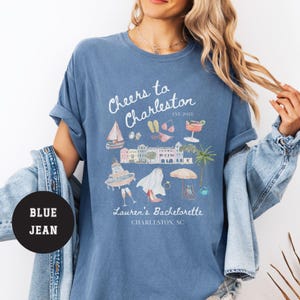K&ouml;nnte beinhalten: Ein blaues T-Shirt mit einem wei&szlig;en Grafikdesign, auf dem "Cheers to Charleston Est. 2025" steht, mit Illustrationen eines Bootes, einer Sonnenbrille, Flip-Flops, eines Cocktails und eines Geb&auml;udes. Der Text "Lauren's Bachelorette Charleston, SC" befindet sich ebenfalls auf dem Shirt.