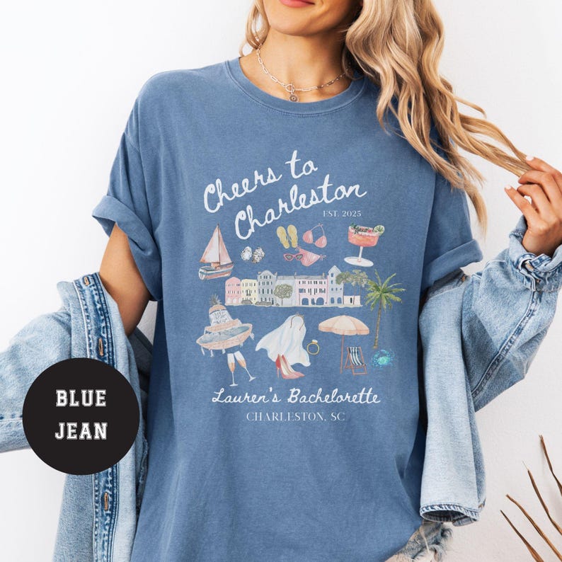 K&ouml;nnte beinhalten: Ein blaues T-Shirt mit einem wei&szlig;en Grafikdesign, auf dem "Cheers to Charleston Est. 2025" steht, mit Illustrationen eines Bootes, einer Sonnenbrille, Flip-Flops, eines Cocktails und eines Geb&auml;udes. Der Text "Lauren's Bachelorette Charleston, SC" befindet sich ebenfalls auf dem Shirt.