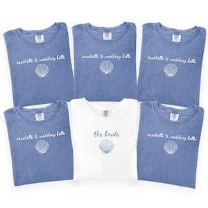 May include: Six t-shirts, five in blue and one in white, are displayed. The blue shirts have the text "seashells & wedding bells" and a seashell graphic. The white shirt says "the bride" with a seashell graphic.