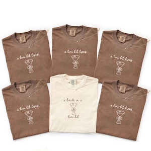 Könnte beinhalten: Sechs T-Shirts werden gezeigt, fünf in Braun und eines in Creme. Die braunen Shirts zeigen den Text "a tire bit tipsy" mit einem Martini-Glas-Design. Das cremefarbene Shirt trägt die Aufschrift "a bride in a tini bit". Jedes Shirt hat eine Halskette.