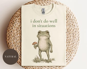 May include: A cream-coloured t-shirt featuring the text "i don't do well in situations" and a cartoon frog holding a mushroom. The frog is green and grey, standing on grass. The shirt is folded on a woven surface.
